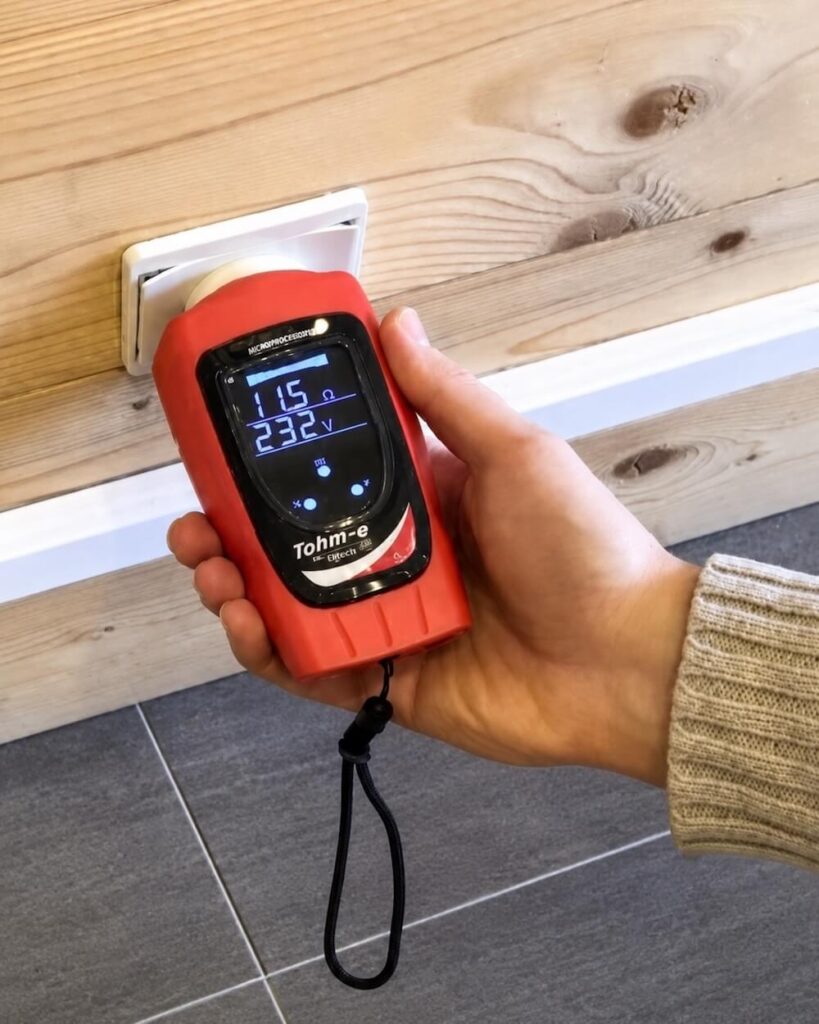 Main tenant un détecteur Tohm‑e T2‑FR100 mesurant l’électricité d’un mur avec prise, affichant la lecture sur l’écran numérique